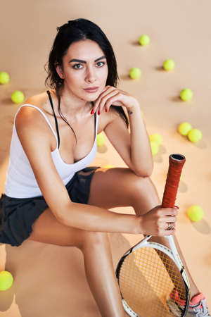 Portrait of beautiful young woman sitting and looking at camera at tennis court among tennis balls and resting and posing dressed in stylish sportswear.の写真素材