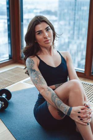 Portrait of skinny brunette girl in stylish sports clothes sitting near window on floor around sport equipment in the background business center.の写真素材