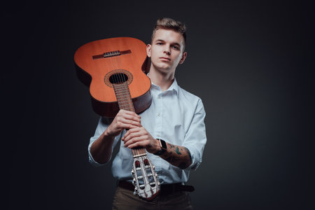 Stylish young dude wearing formal suit poses in dark background holding acoustic guitar. Attractive man with fashionable hairstyle.の写真素材