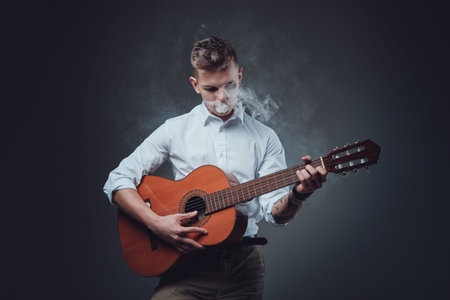 Portrait of a hipster guy dressed in formal clothing playing guitar in dark background and smoking...