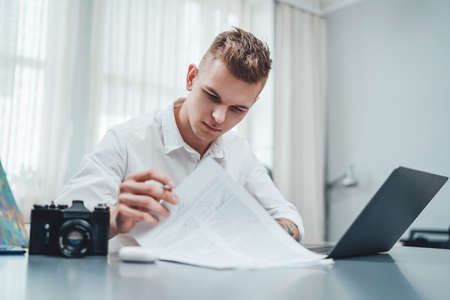 Career and business. Portrait of a thoughtful man dressed in shirt and doing his paperwork sitting at table in modern office room.の写真素材