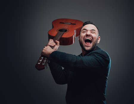 Savage bearded man poses in dark background holding acoustic guitar and screaming to camera. Angry guy dressed in stylish suit.の写真素材