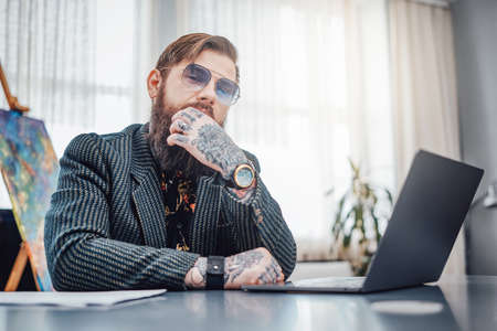 Work at home. Man dressed in fashionable suit with sunglasses sits at table with a laptop. Brutal guy and his remote work.の写真素材