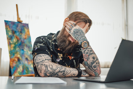 Stylish guy with tired arms sits at table doing his job at home. Man and his remote work he does it...