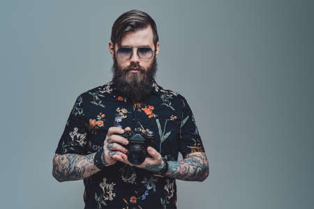 Successful hipster person with bearded face and modern coiffure posing in gray background holding photo camera.の写真素材