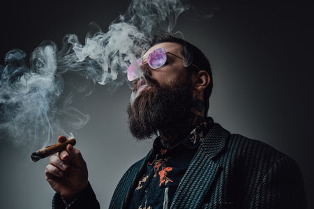 Elegant man in retro style poses in dark background smoking cigar. Portrait of a bearded guy with stylish coiffure.の写真素材