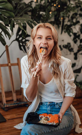 Joyful blond eating sushi sitting on table around houseplantsの写真素材