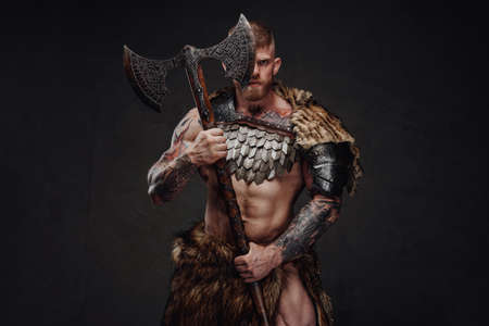 Brutal tattooed warrior wearing light armour and fur holding two-handed axe in dark studioの写真素材
