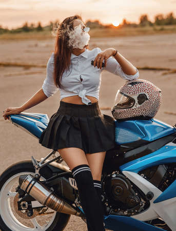 Fashionable girl biker smoking vape leaning motorcycle at sunset