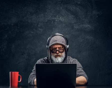Tricky elderly man with hood and headphones using laptopの写真素材