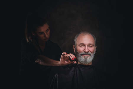 Female barber with scissors cutting hairs of senior manの写真素材