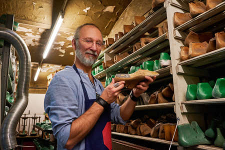 Elderly man with apron holding shoe around shelfs with boots