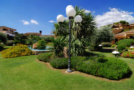 House/hotel in the beautiful summer garden with many plants and path - blue sky with clouds above and a lamp in the frontの写真素材