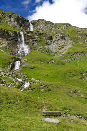 Beautiful alpine waterfall going down directly from the peaks through green  meadowsの写真素材