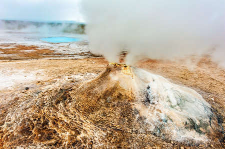 Fumarole in the geothermal area Hveravellir, central Iceland. The area around is layered and cracked. In back is visible incredibly blue silica pool Blahverの写真素材