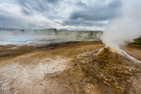 Fumarole in the geothermal area Hveravellir, central Iceland. The area around is layered and cracked. In back is visible incredibly blue silica pool Blahverの写真素材