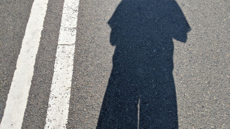 The shadow of a person standing on a paved road. The person is wearing pants and a shirt, and their arms are at their sides. A white line runs down the center of the road.の写真素材