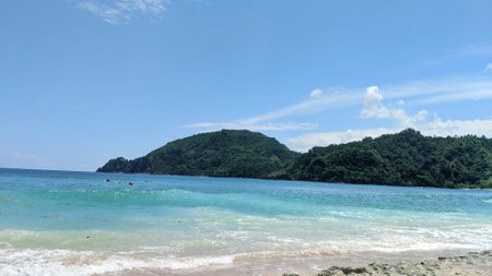 Turquoise waves meet a sandy beach with lush green hills in the background under a blue sky with...