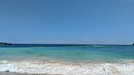 Turquoise waves crash onto a sandy beach under a clear blue sky. The horizon line divides the sea and sky in this serene coastal scene. Wediombo Beach Gunungkidul, Yogyakarta, Indonesia.の写真素材