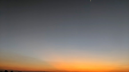 A vibrant sunrise sky with a gradient of deep orange at the horizon transitioning to soft blue-gray at the top, featuring a single bright star or planet.の写真素材