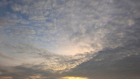 A dramatic sky filled with a mosaic of small, textured clouds, predominantly grey with patches of warm orange and yellow light peeking through, hinting at a sunrise or sunset.の写真素材