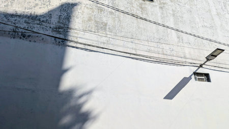 A minimalist urban scene featuring a textured white wall, stark shadows, power lines, and a street light, capturing architectural details and graphic elements.の写真素材
