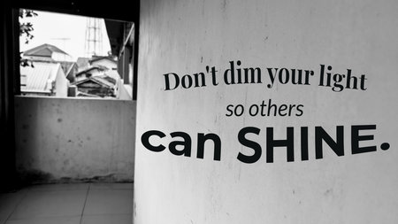A black and white photo of a wall with a window, displaying the quote, Don&#39;t dim your light so others can SHINE.の写真素材