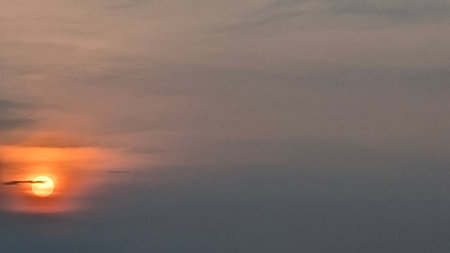 A beautiful sunrise scene with the sun shining brightly through thin clouds, creating a striking orange glow. The sky transitions from a deep gray to warm, soft colors near the horizon.の写真素材