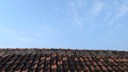 A clear blue sky with a few wispy clouds is partially covered by the dark, weathered roof tiles of a...
