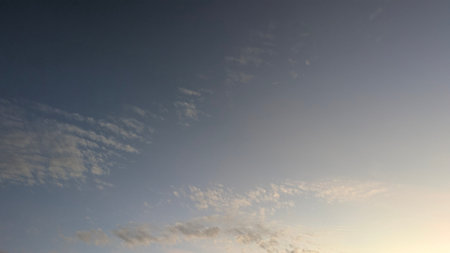 A tranquil sky at dawn. The top half is a rich, dark blue while the bottom fades to a soft, hazy peach. Wispy and scattered clouds drift through the peaceful sunrise atmosphere.の写真素材