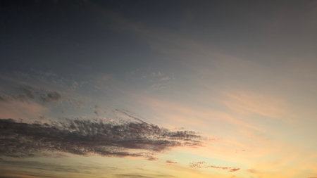 A tranquil sunrise sky shows a gradient from dark blue to a soft, peach-colored horizon. There are different types of clouds from thin streaks to more textured, dramatic formations.の写真素材