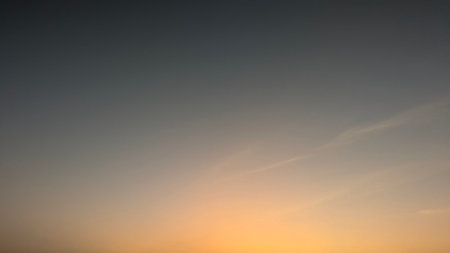 A serene sunrise sky with a soft gradient of orange and yellow at the bottom, blending into a deep blue-gray above. Two small, faint dots are visible in the distance.の写真素材