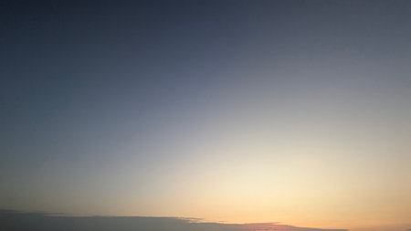 A serene image of a pale blue sky. The top of the image is a solid dark blue, which smoothly transitions to a soft white and orange glow at the bottom. A thin, dark cloud is visible at the very bottomの写真素材