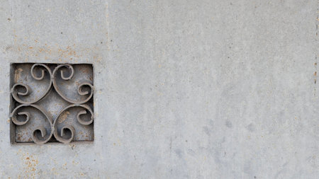 A detailed close-up shot of a grey wall shows a small square metal grating with an elegant swirl pattern. The surface of the wall appears textured and slightly weathered with small rust stains.の写真素材