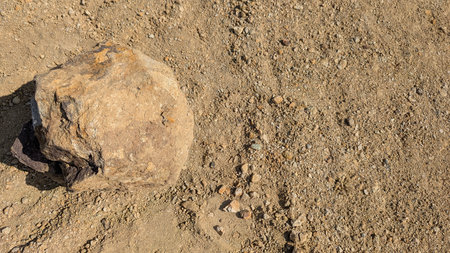 A large, weathered boulder rests on a sandy, tan-colored ground. The surface is covered in fine grit...