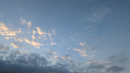 A dawn sky with a striking contrast. The lower section features dark, heavy clouds, while the upper-left sky has scattered, sunlit golden clouds against a soft blue background.の写真素材