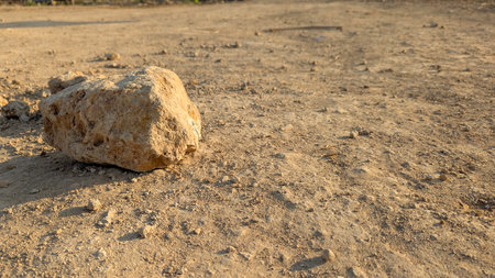 A close-up of a rugged, sandy landscape. The ground is a mix of fine sand, small rocks and pebbles. A prominent, large rock is in the foreground, casting a shadow, creating a textured, natural surfaceの写真素材