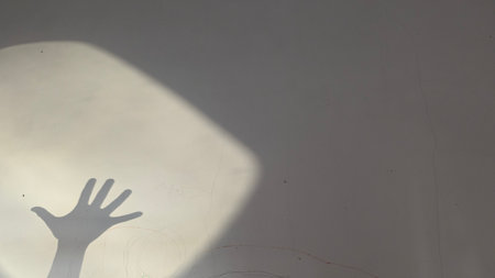 A dramatic shadow of an outstretched human hand projected onto a textured wall, partially illuminated by a bright, diagonal spotlight effect creating a high-contrast, moody, and abstract compositionの写真素材