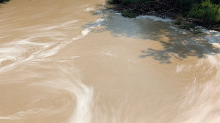 An artistic long exposure of a muddy brown river, showing silky white swirls of moving water, with a...