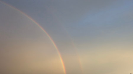 A full double rainbow arches across a vast, twilight sky. Muted blue, gray, soft yellow, orange light dominate the vertical frame, creating an ethereal, serene, atmospheric texture.の写真素材