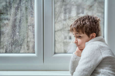 portrait of a pensive young man looking away through a wet window in a rainy day. Toned image.の写真素材