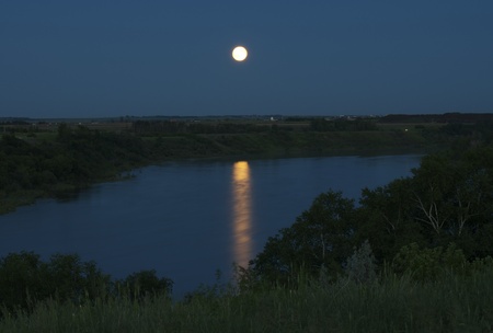 The glowing full moon is bright against the dark blue sky  Reflecting on the South Saskatchewan River  The water looks very smooth and there is green grass in the foreground with the valleyの写真素材