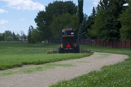 In the park area a large riding lawnmower is cutting grass next to the walking trail, the machine is moving away from the camera.の写真素材