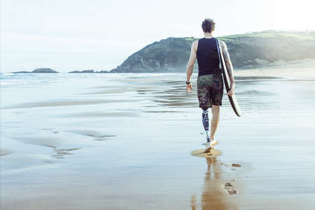 Caucasian surfer man, seen from behind, walking along the beach with an artificial leg.Copy Spaceの写真素材