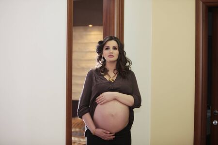 Pregnant attractive young woman with hand over her belly looking at the camera inside her homeの写真素材