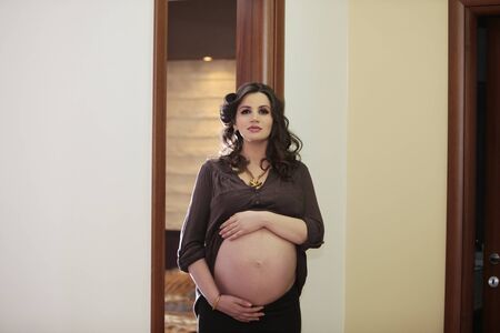 Pregnant attractive young woman with hand over her belly looking at the camera inside her homeの素材 [FY3108852952]