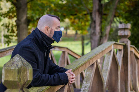 A man leaning on the railing of an old wooden bridge in a local park in maskの写真素材