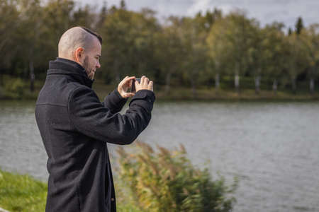 A man takes a picture at a small lake with his mobile phoneの写真素材