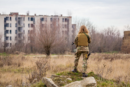 A beautiful girl in military uniform with an airsoft gun standing on a mossy stone heap with ruined buildings in the backgroundの写真素材