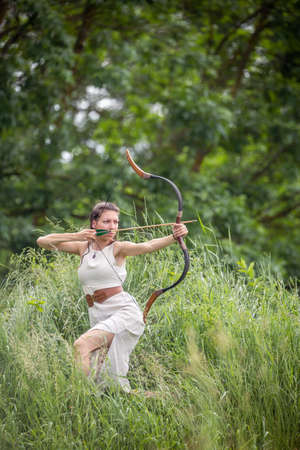 A Hungarian woman in a linen dress kneeling with a bow in the tall grassの写真素材